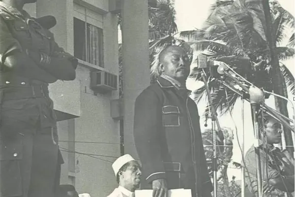 Historical black-and-white photograph of Comorian revolutionary leader Ali Soilih delivering a speech at outdoor microphones, accompanied by armed soldiers and government officials.