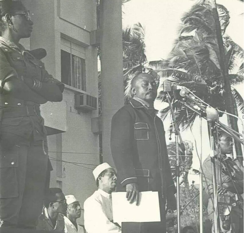 Historical black-and-white photograph of Comorian revolutionary leader Ali Soilih delivering a speech at outdoor microphones, accompanied by armed soldiers and government officials.