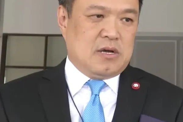 A man in a dark suit and blue tie speaking into microphones
