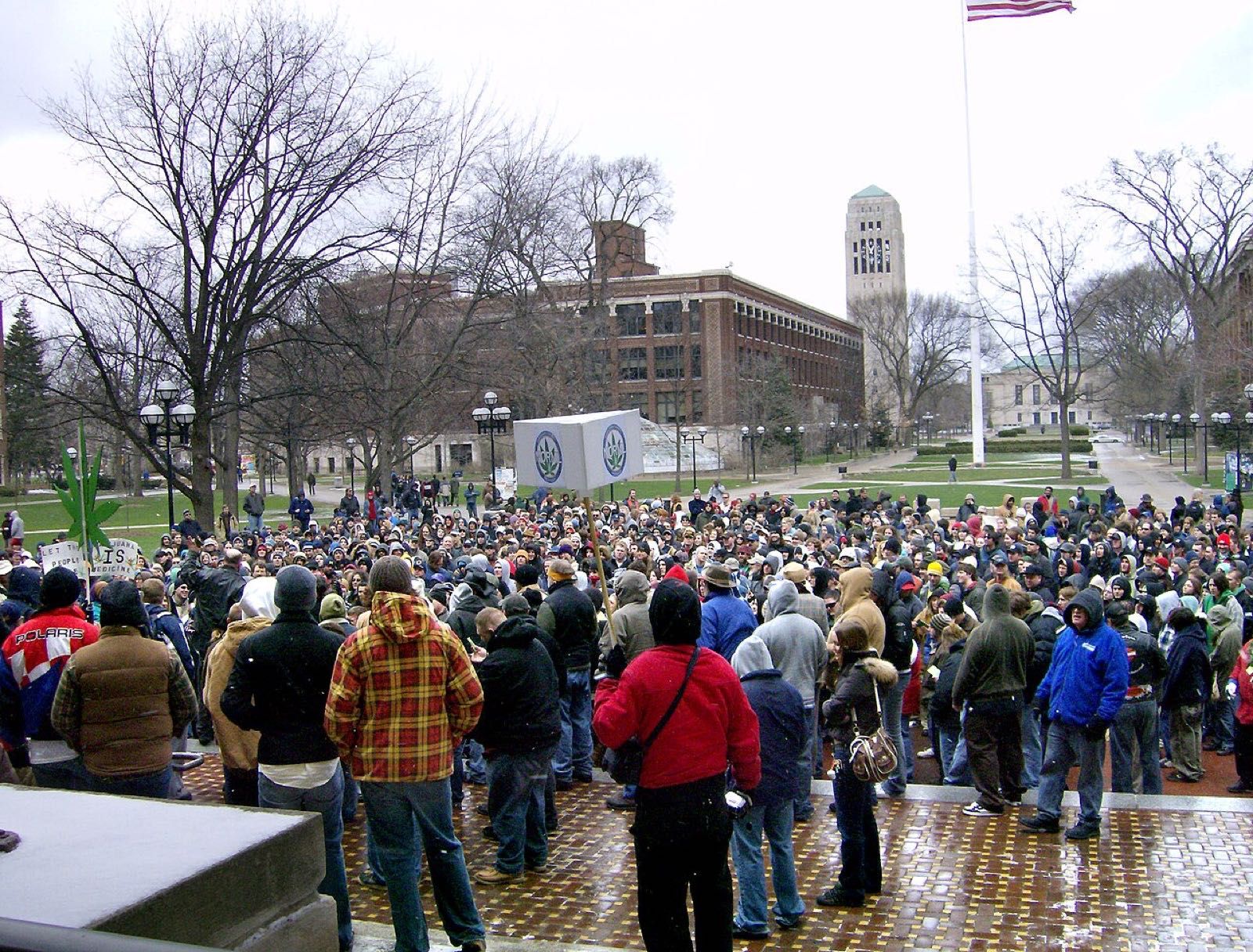 A crowd on a university square around a speaker and a large model of a cannabis leaf on a pole