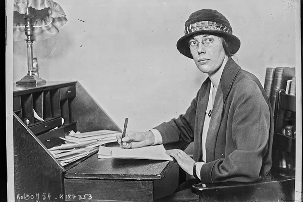 Black-and-white press photograph of a woman in a hat and coat writing at a desk