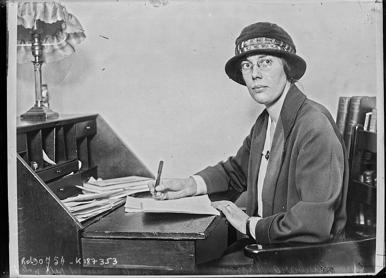 Black-and-white press photograph of a woman in a hat and coat writing at a desk