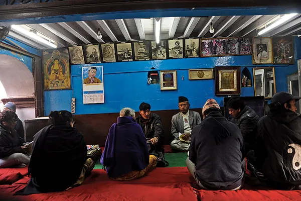 Documentary color photograph of Nepali men seated together on floor mats inside a Kathmandu devotional gathering space, with religious pictures on the walls.