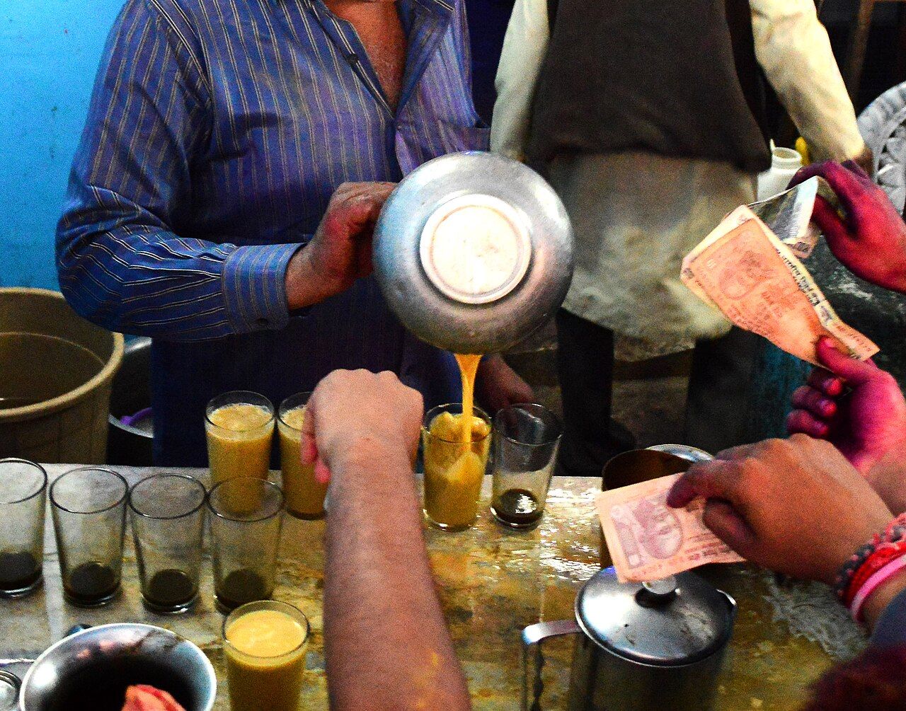 A man pours thandai from a metal pot into a row of glasses while customers hold out banknotes