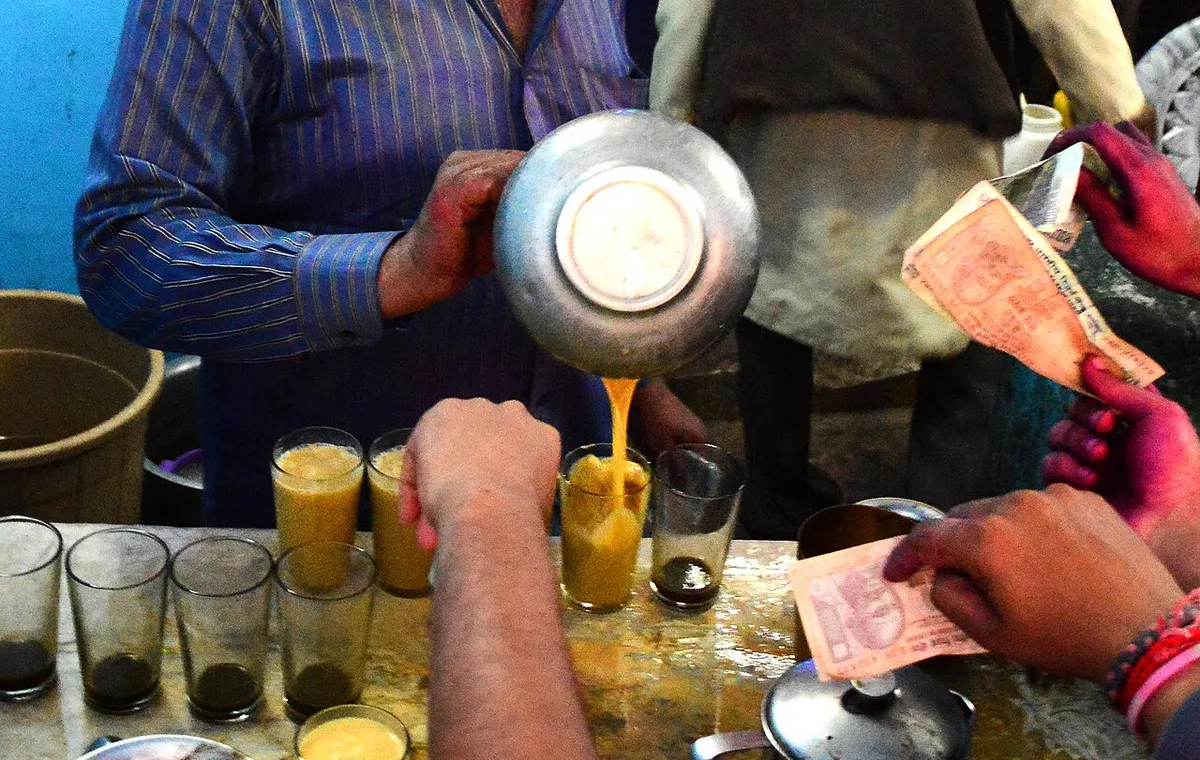 A man pours thandai from a metal pot into a row of glasses while customers hold out banknotes