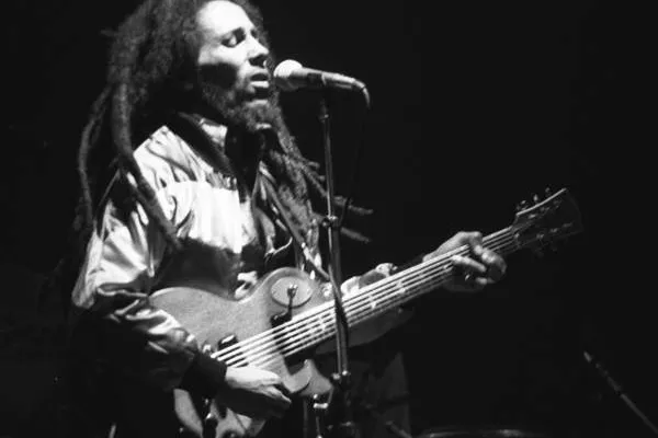 A man with long dreadlocks singing into a microphone and playing an electric guitar on a dark stage