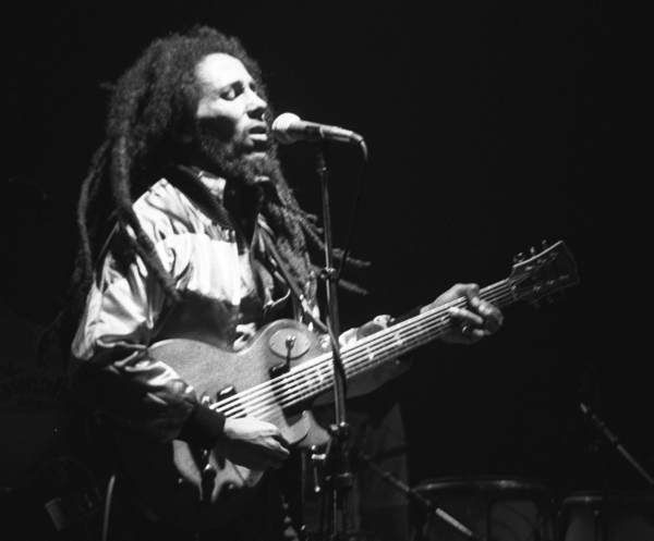 A man with long dreadlocks singing into a microphone and playing an electric guitar on a dark stage