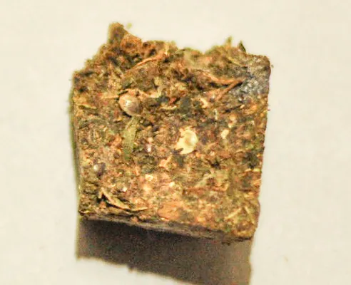 A compact square block of pressed cannabis with visible leaf fragments and seeds on a pale background.