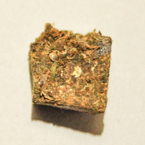 A compact square block of pressed cannabis with visible leaf fragments and seeds on a pale background.