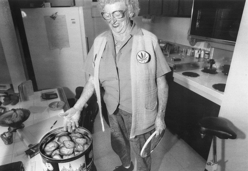 Black-and-white photograph of Mary Jane Rathbun in a kitchen, wearing glasses and a cannabis-leaf badge beside a tin of baked goods.