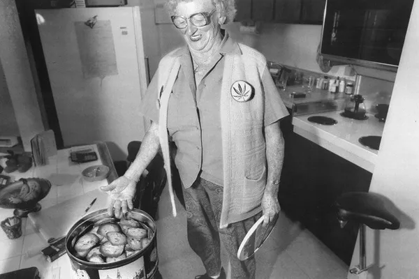 Black-and-white photograph of Mary Jane Rathbun in a kitchen, wearing glasses and a cannabis-leaf badge beside a tin of baked goods.