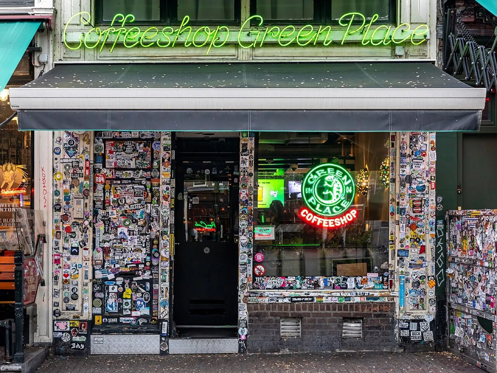 The front of an Amsterdam coffeeshop, its windows covered in stickers and lit by green neon