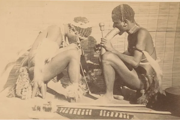 Sepia photograph of two seated African men; the one on the right inhales from a large horn water pipe, the one on the left bends low over a long thin reed reaching to the floor.