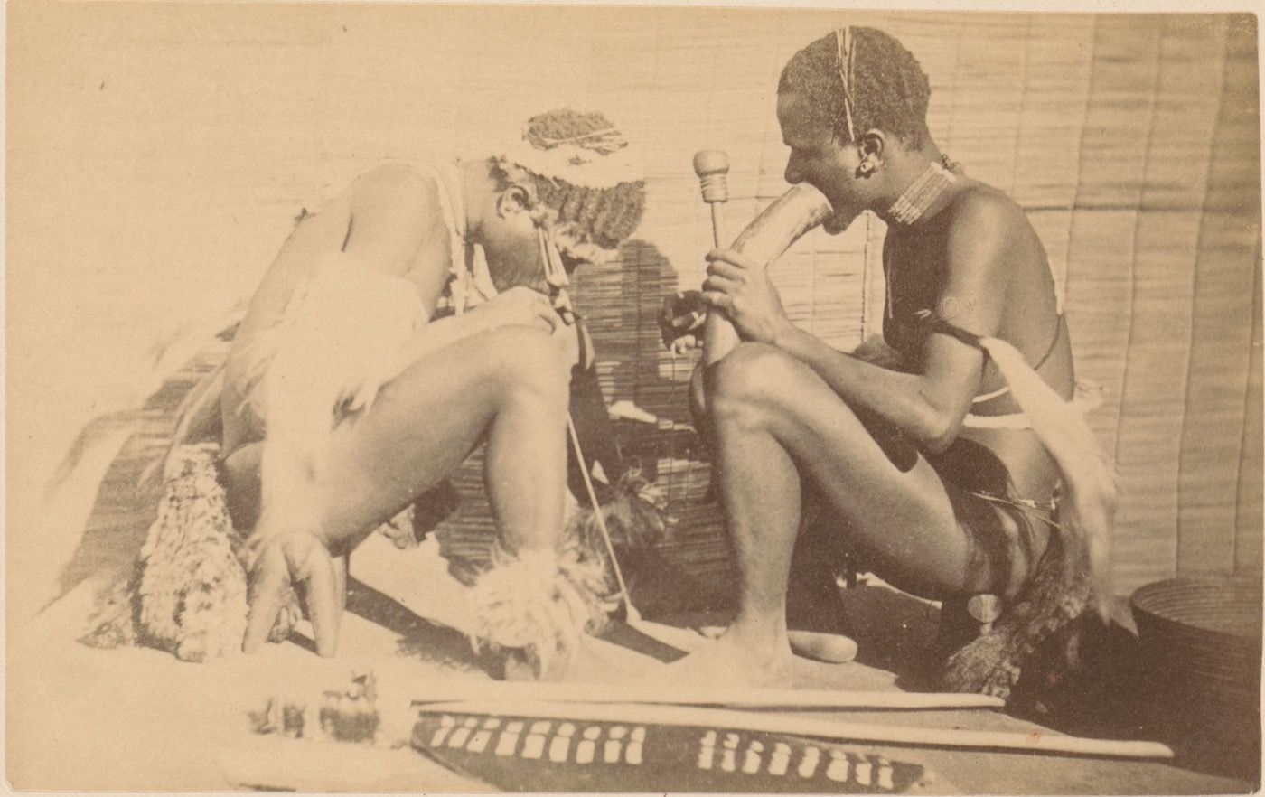 Sepia photograph of two seated African men; the one on the right inhales from a large horn water pipe, the one on the left bends low over a long thin reed reaching to the floor.