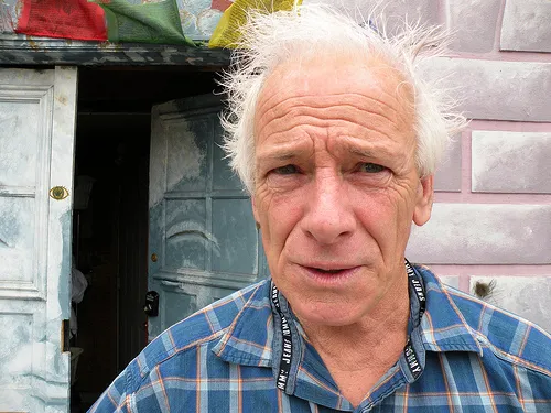Dennis Peron with white hair wearing a blue checked shirt outside a doorway.