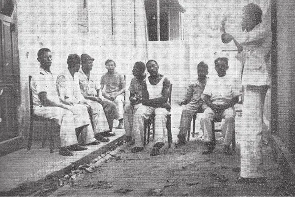 Historical black-and-white photograph of a group gathered in a Brazilian courtyard, with seated participants and one person standing at the right, reproduced as a clube de diambistas in Propriá, Sergipe.