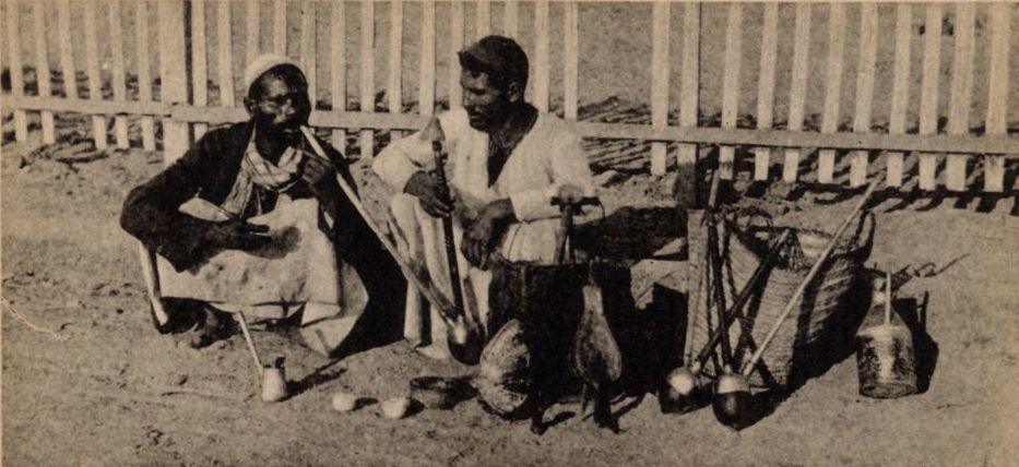 An old postcard of two men smoking long pipes in front of a white fence