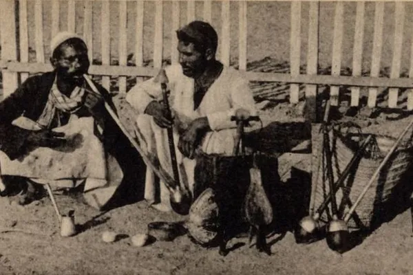 An old postcard of two men smoking long pipes in front of a white fence