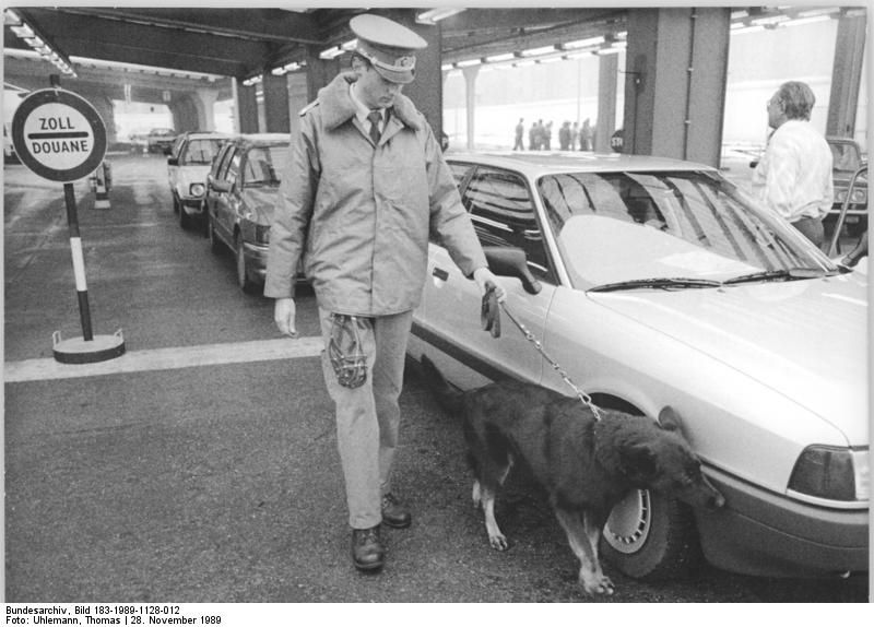 Black-and-white photograph of an East German customs officer with a drug-detection dog inspecting a car at Berlin's Heinrich-Heine-Straße border crossing in November 1989.
