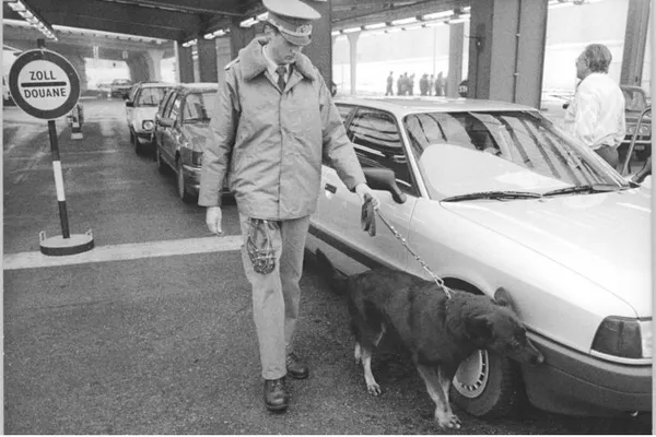 Black-and-white photograph of an East German customs officer with a drug-detection dog inspecting a car at Berlin's Heinrich-Heine-Straße border crossing in November 1989.