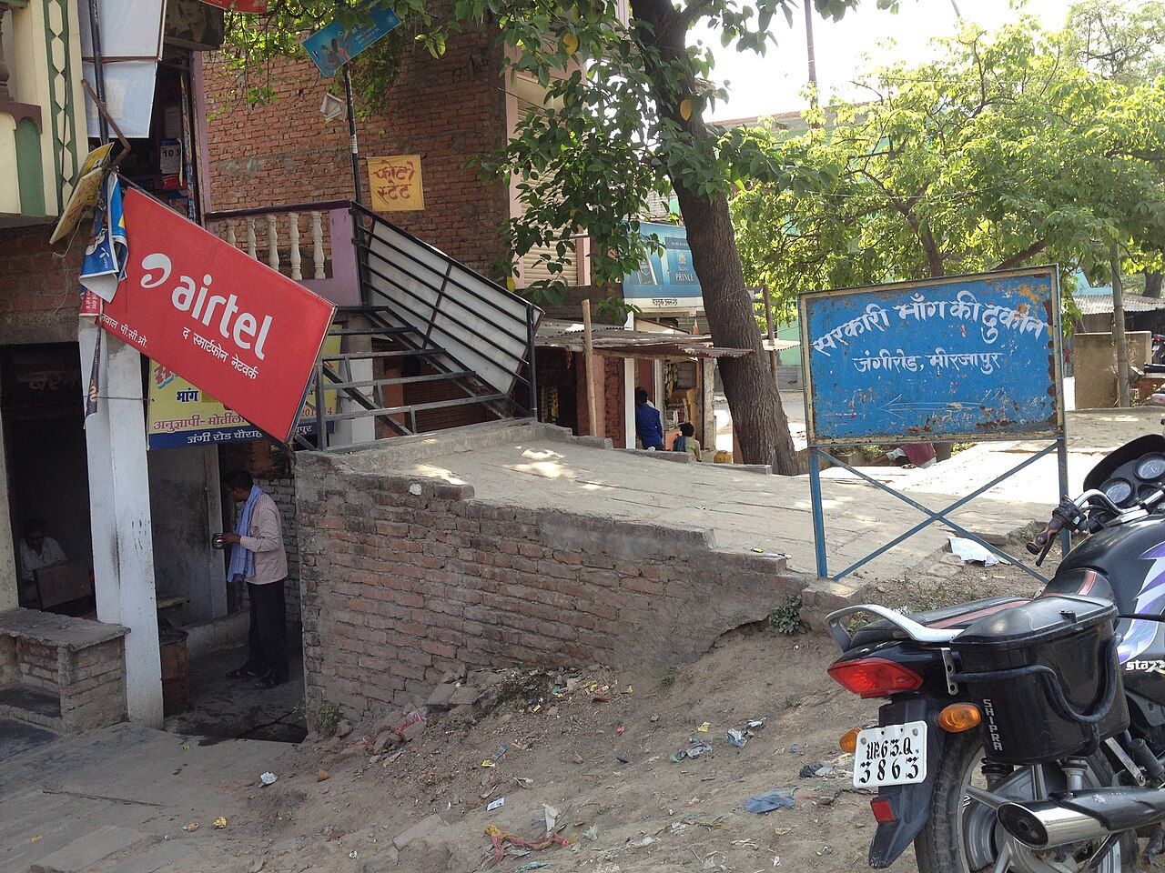A street corner with a blue roadside sign in Hindi reading "government bhang shop, Jangi Road, Mirzapur", and a small yellow sign reading "bhang" over a shop doorway.