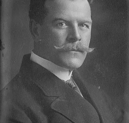 Black-and-white studio portrait of a man with a large moustache in a dark suit and tie