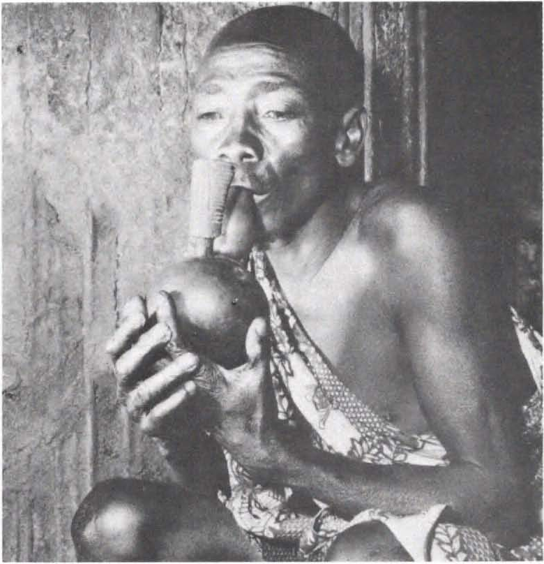 Historical black-and-white ethnographic photograph of a Twa man in Rwanda holding a traditional gourd water pipe, used in anthropological interviews about cannabis in 1959–60.