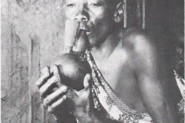 Historical black-and-white ethnographic photograph of a Twa man in Rwanda holding a traditional gourd water pipe, used in anthropological interviews about cannabis in 1959–60.