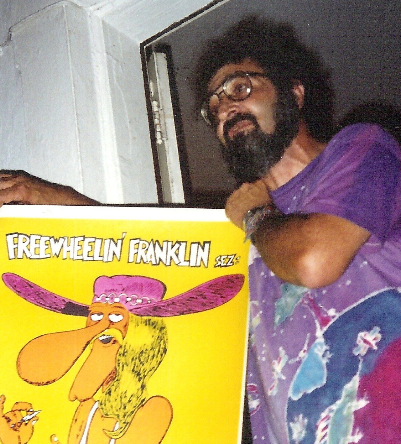 A bearded man in glasses and a tie-dyed shirt leaning out beside a bright comic-book poster