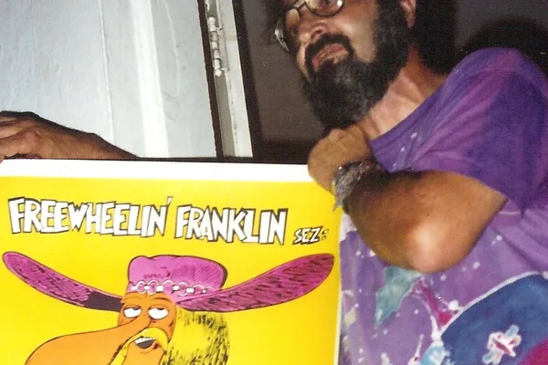 A bearded man in glasses and a tie-dyed shirt leaning out beside a bright comic-book poster