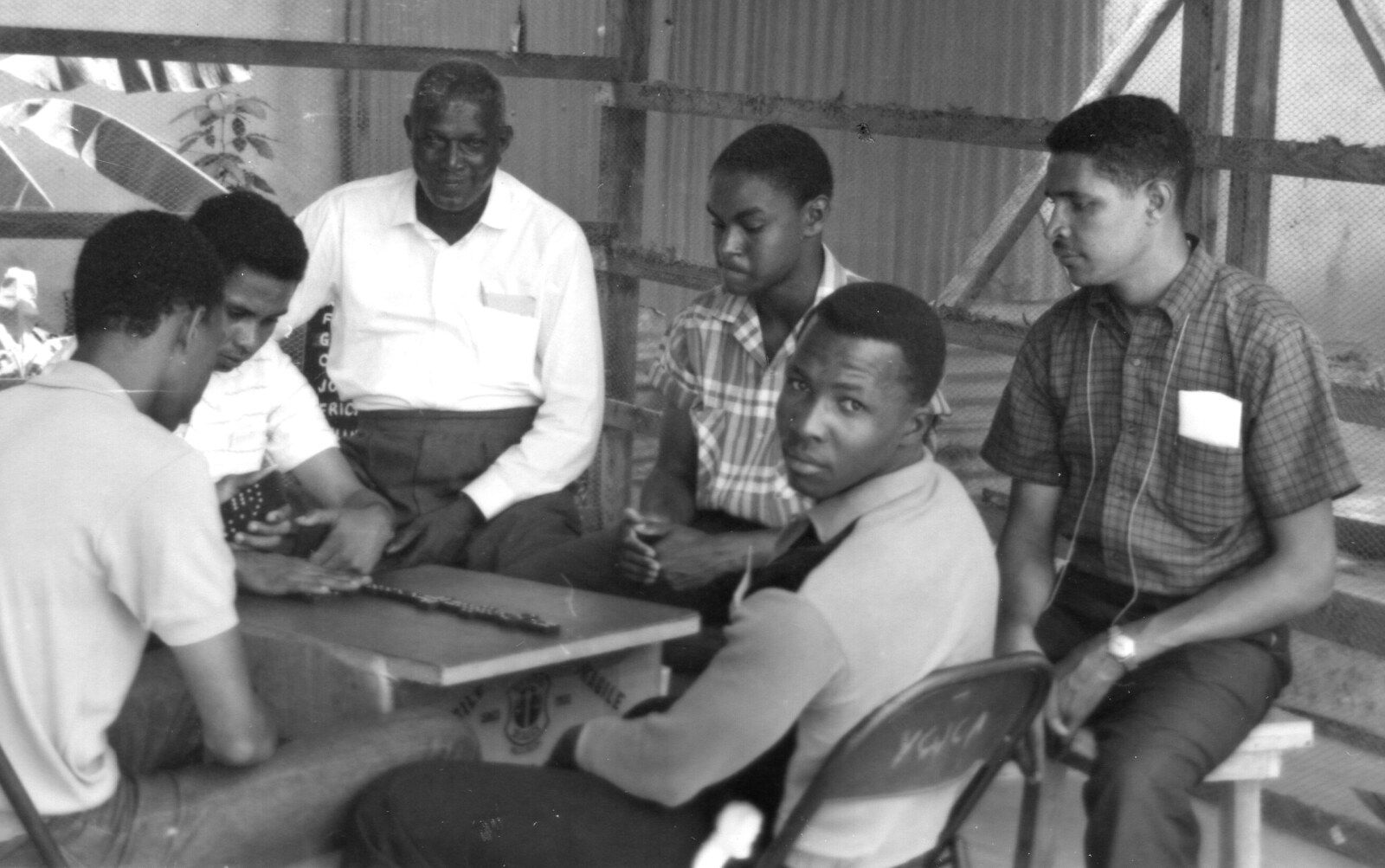 Historical black-and-white photograph of Jamaican men gathered around a domino table in a modest wooden shelter in 1964.