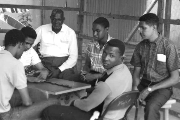 Historical black-and-white photograph of Jamaican men gathered around a domino table in a modest wooden shelter in 1964.