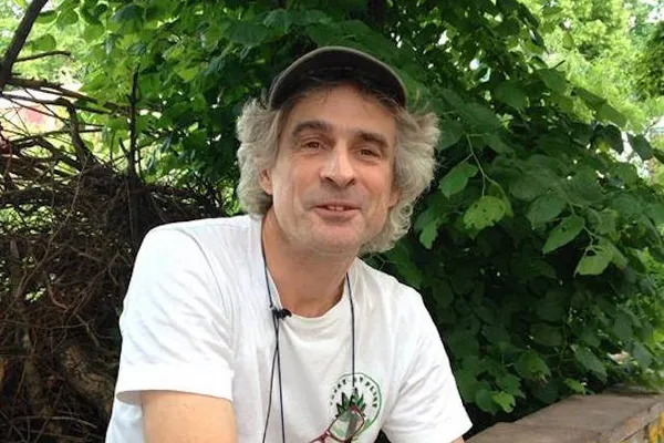 Joep Oomen sitting outdoors in front of green vegetation, wearing a white Trekt Uw Plant T-shirt and a dark cap.