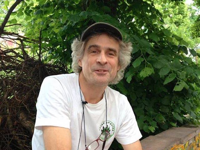 Joep Oomen sitting outdoors in front of green vegetation, wearing a white Trekt Uw Plant T-shirt and a dark cap.