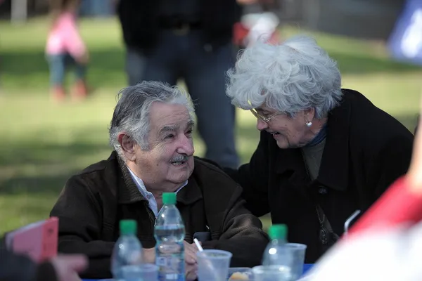 An elderly man in a dark jacket sits at an outdoor table, smiling, while a woman beside him leans in to speak