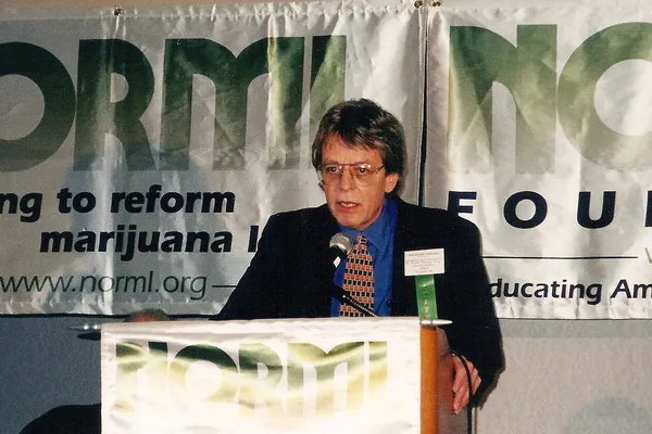 A man in glasses and a dark jacket speaking at a lectern in front of large NORML banners