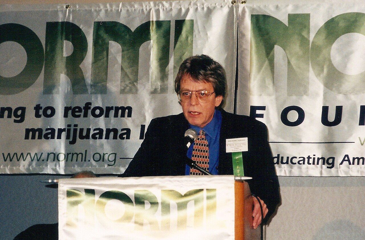 A man in glasses and a dark jacket speaking at a lectern in front of large NORML banners