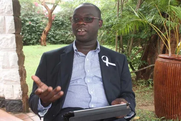 Kenyan parliamentarian Ken Okoth seated outdoors wearing glasses and a dark blazer, gesturing with one hand while holding a tablet.
