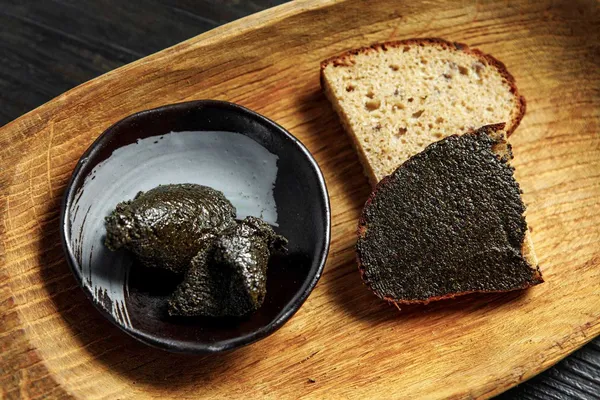 Traditional Latvian dark hemp-seed butter in a ceramic bowl beside rustic rye bread on a wooden board.