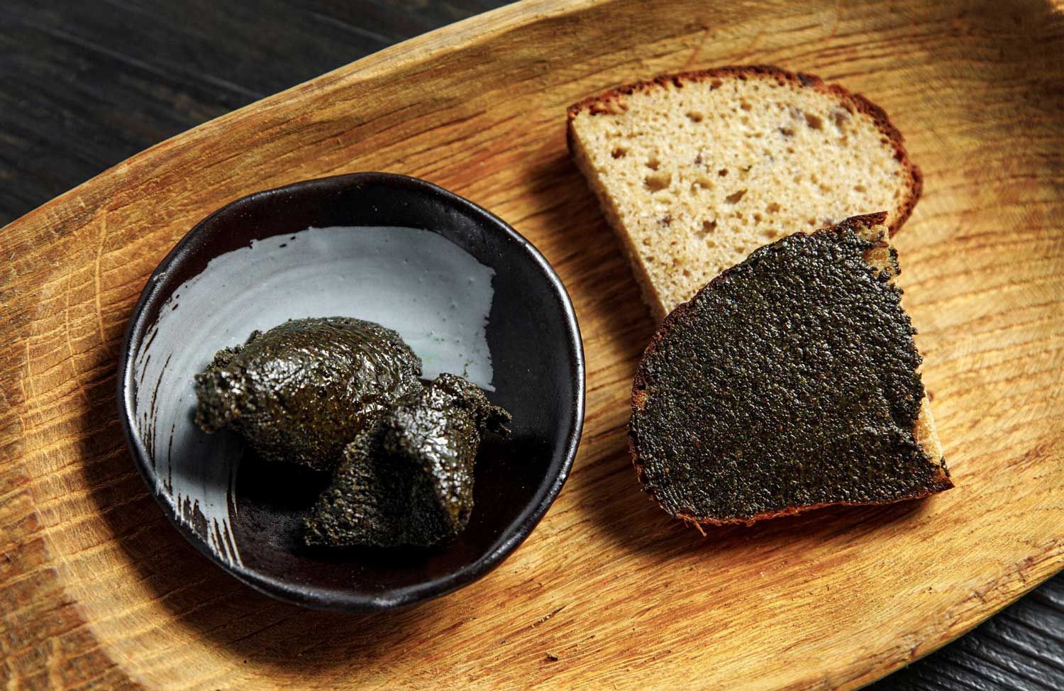 Traditional Latvian dark hemp-seed butter in a ceramic bowl beside rustic rye bread on a wooden board.