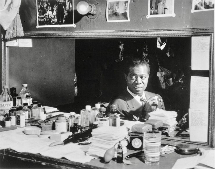 Louis Armstrong in a suit sitting at a cluttered dressing-room table under a mirror and lights