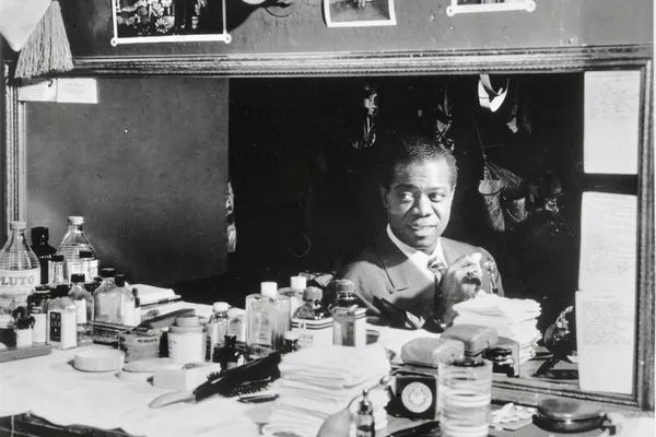 Louis Armstrong in a suit sitting at a cluttered dressing-room table under a mirror and lights