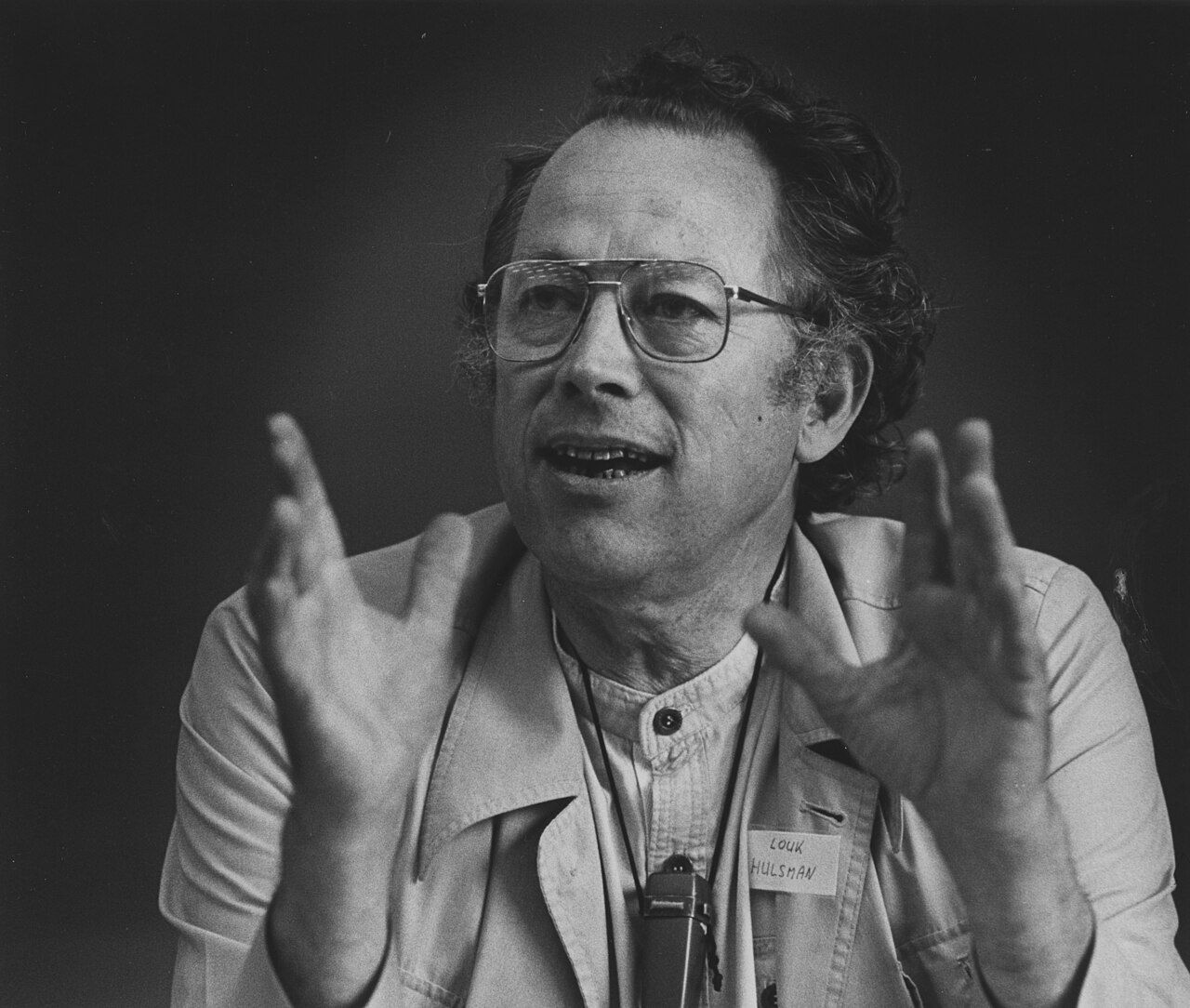 Black-and-white photograph of a man in glasses and a light jacket speaking with both hands raised, a name tag on his chest