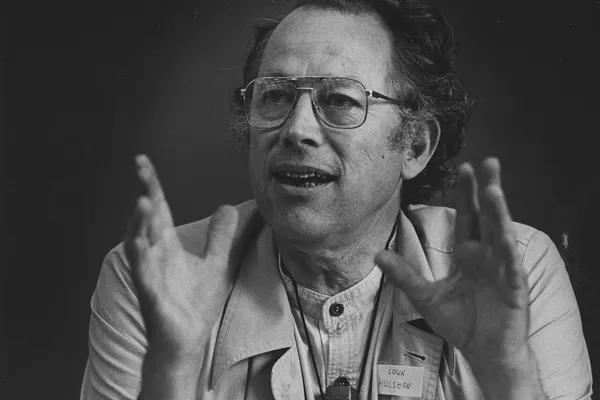 Black-and-white photograph of a man in glasses and a light jacket speaking with both hands raised, a name tag on his chest