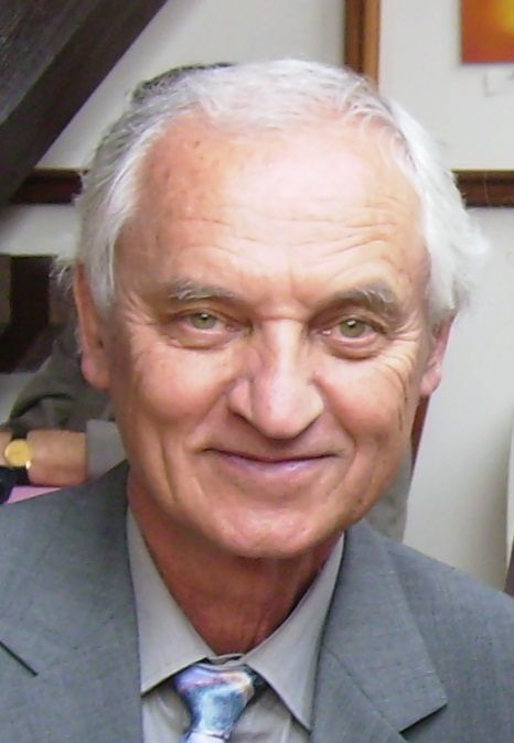 A smiling white-haired man in a grey suit and patterned tie, photographed indoors