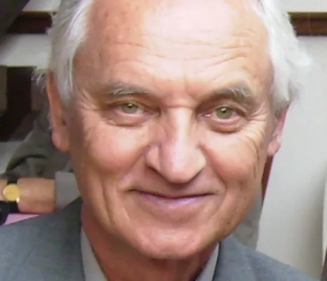 A smiling white-haired man in a grey suit and patterned tie, photographed indoors