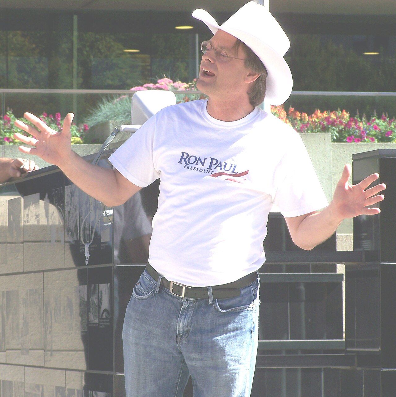 A man in glasses, a white cowboy hat and a white T-shirt speaking with his arms spread at an outdoor rally