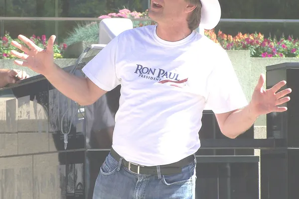 A man in glasses, a white cowboy hat and a white T-shirt speaking with his arms spread at an outdoor rally