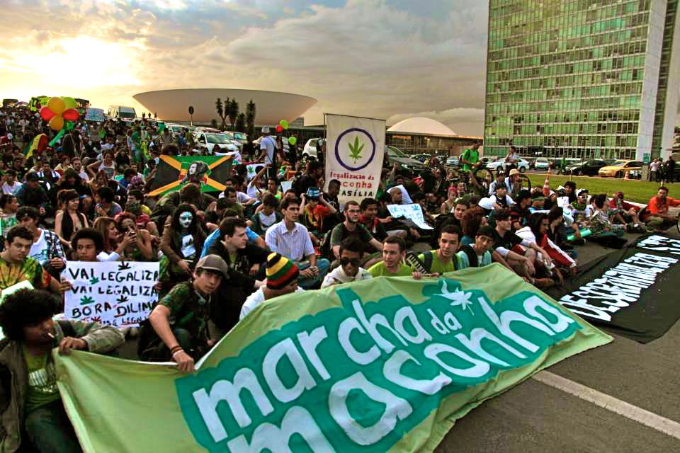 Young people sitting on a road behind a large green "marcha da maconha" banner, with Brazil's National Congress building behind them.