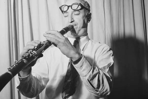 Black-and-white photograph of a man in shirt sleeves and tie playing a clarinet, eyes closed, glasses pushed up on his forehead
